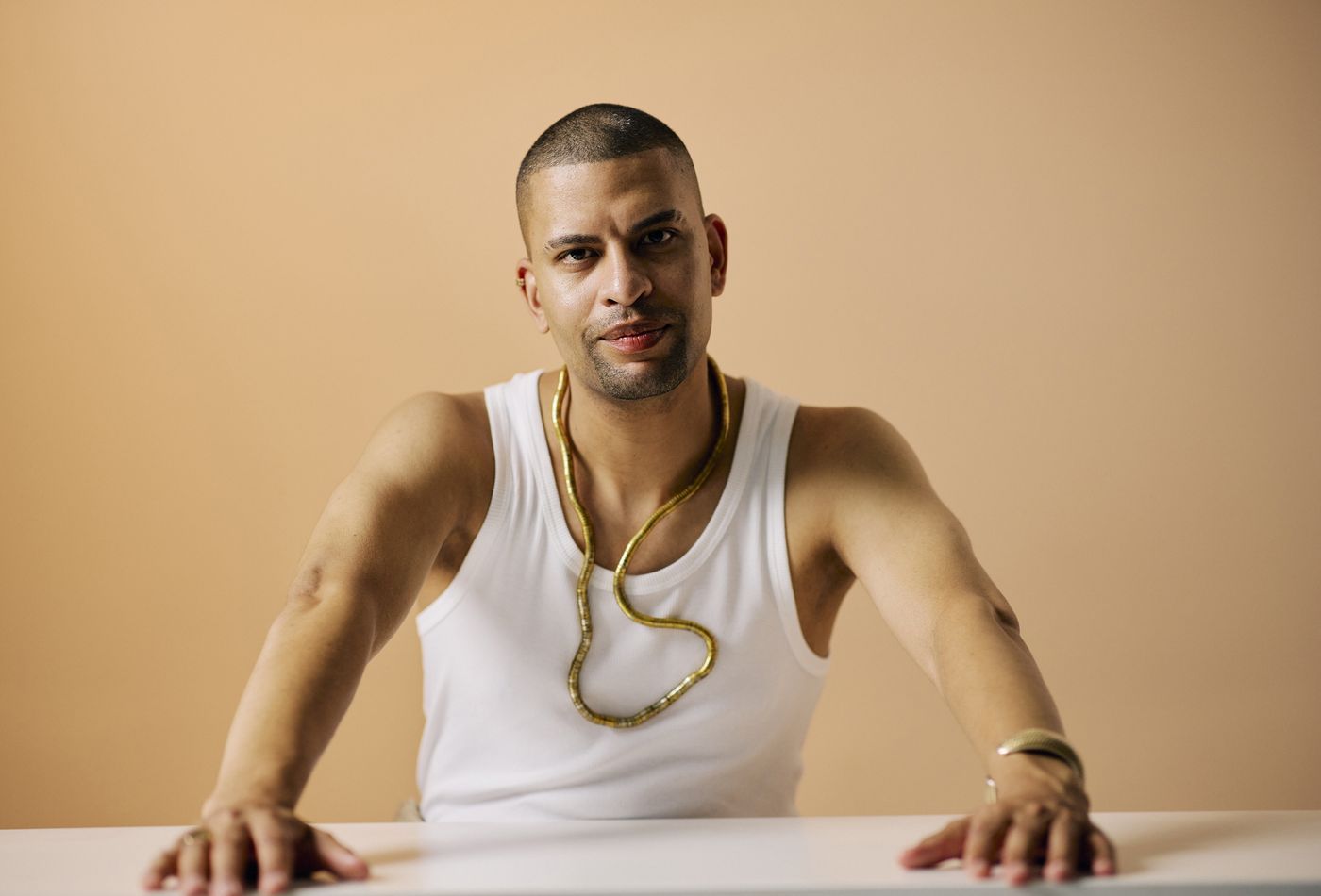 Traidy Naidoo at a table in a white vest and a gold chain, hands resting on the surface, looking straight at the camera.