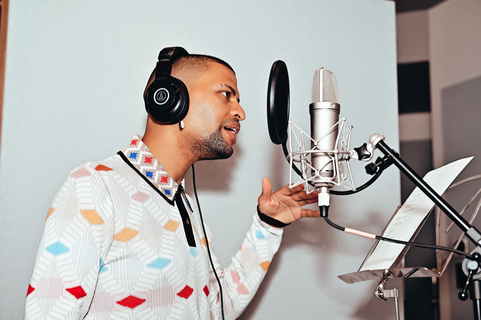 Traidy Naidoo in profile at a studio microphone, headphones on, mouth open mid-phrase with one hand raised.
